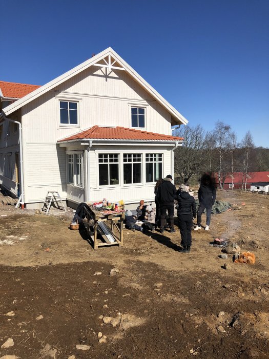 Personer grillar och socialiserar utanför ett nybyggt vitt hus med en oarbetsam tomt i soligt väder.