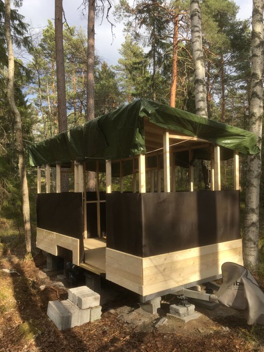 Under uppbyggnad av träramverk för ett litet hus med reglar, takstolar och delvis täckt med grön presenning.