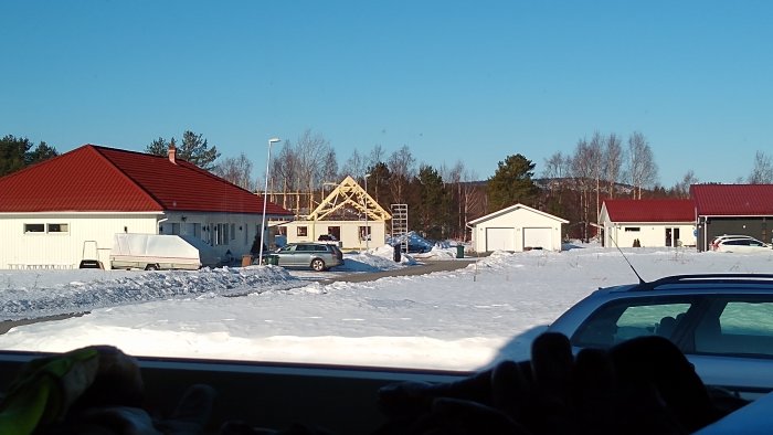 Vy genom ett snöigt fönster på ett pågående byggprojekt av ett trästomme hus, med andra hus och en bil i förgrunden.