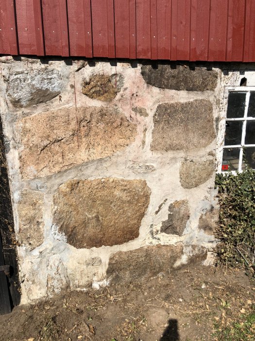 Delvis målad stenmur med synliga fogar och omålade stenar vid en röd byggnad.