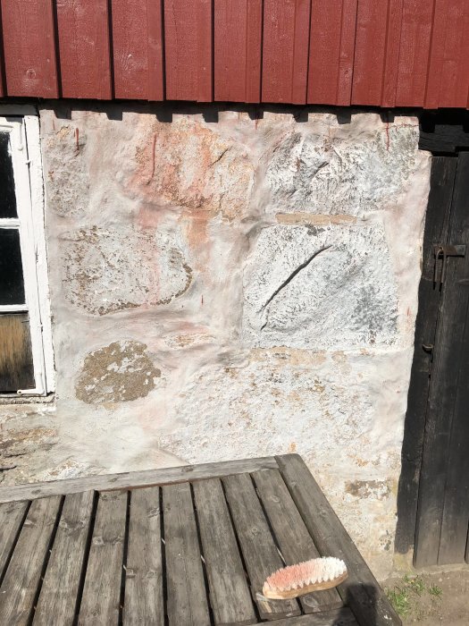 En vägg med ojämn plastfärg bredvid en svart dörr, röda paneler ovanför och en skurborste på en träbänk.