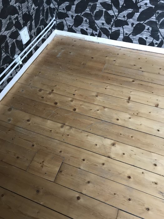 Hörn av ett rum med traditionellt trägolv med synliga nötningar och en vägg med grafiskt bladmönstrad tapet.