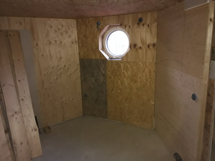 Källare med nyinredda väggar av plywood och OSB-skivor, oinredda golv och ett runt fönster.