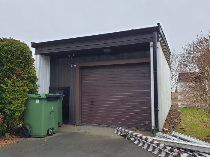 Garage i mörkbrunt med tillhörande soptunnor bredvid en nedtagen takränna.