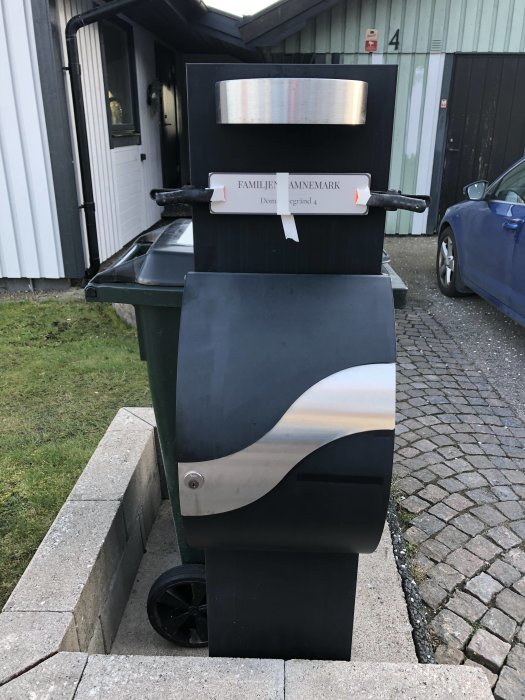 Svart modern brevlåda på ställning med personligt namnskylt framför ett hus.