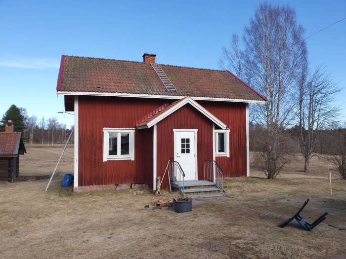 Röd timmerstuga från 1870-talet före renovering, med gräsmatta och blå himmel.