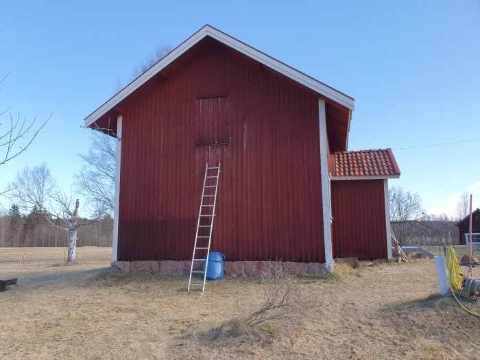 Röd timmerstuga från 1800-talet med takvind, ståltrappa och blå tunna bredvid, innan utbyggnadsprojektet påbörjats.