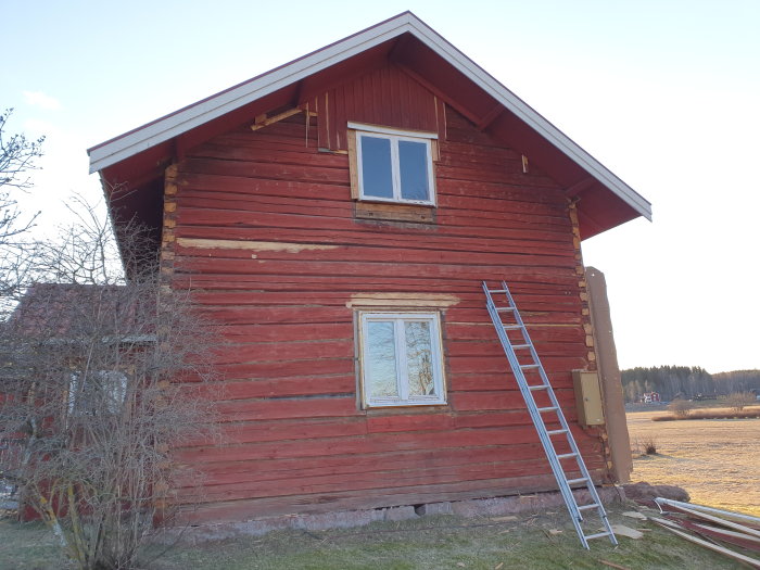 Röd timmerstuga under renovering med borttagen veranda och en stege lutad mot väggen.