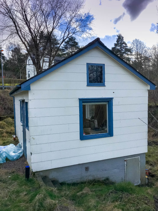 Litet renoveringsobjekt, vitmålat hus med sovloft under lutande blått tak mot naturbakgrund.