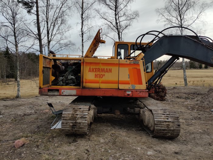 Gul och orange grävmaskin av märket Åkerman på en tom obebyggd tomt med öppen motorlucka och delvis synlig motor.