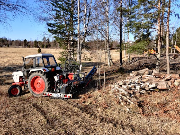 Traktor vid hög med nedsågade trädstammar på en gård, beredda för att göras till ved.