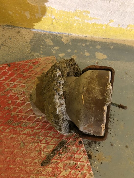 Ett rostat tätningslock ingjutet i betong, borttaget från rör, på ett gult och grått golv med röda spår.