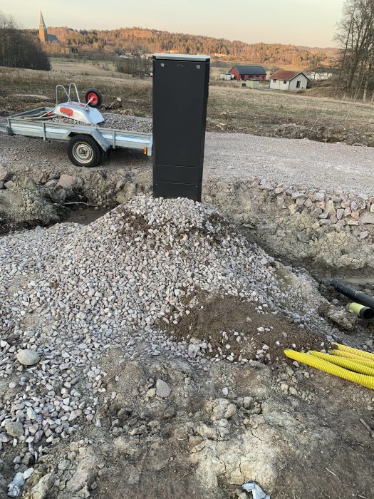 Ett svart elskåp monterat på ett underrede bland bygggrus, med en markgrävning och tomrör i förgrunden och en släpvagn i bakgrunden.