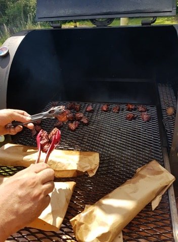 En person vänder köttbitar på en grill med tång, med inslagpapper på sidan.