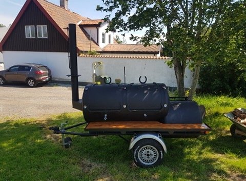 Svart smoker grill på släpvagn framför hus och bil.