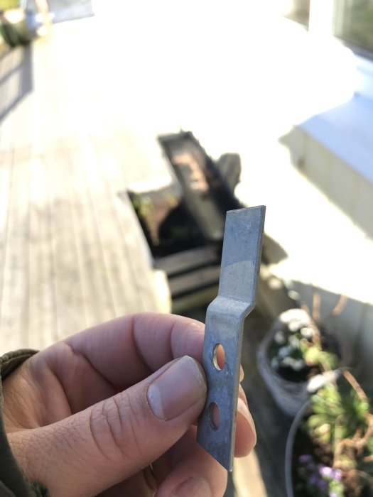 Hand håller ett litet metallbeslag med hål för skruv, mot suddig bakgrund av en trädäck.