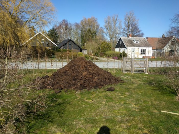 En stor hög med hästgödsel i en trädgård bredvid buskar och ett staket med hus i bakgrunden.