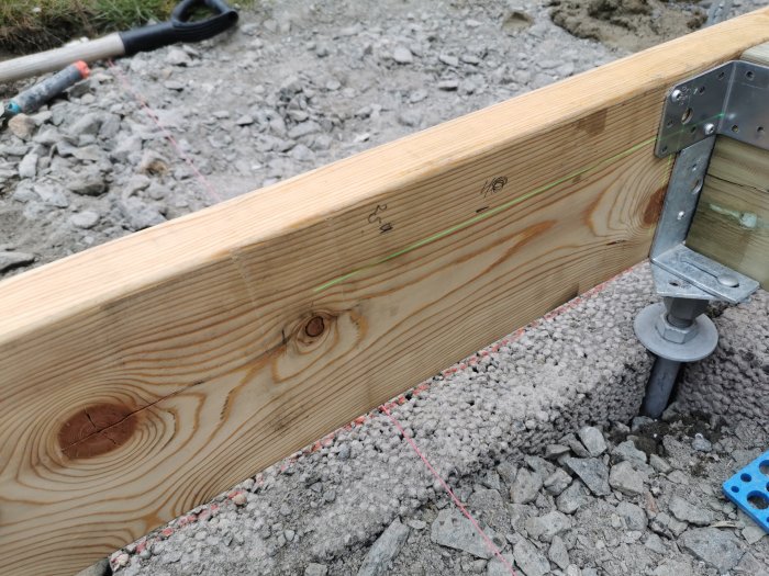 En träregel på marken stabiliserad med en justerbar stödsko, markerad för trappbygge, med grus och verktyg i bakgrunden.