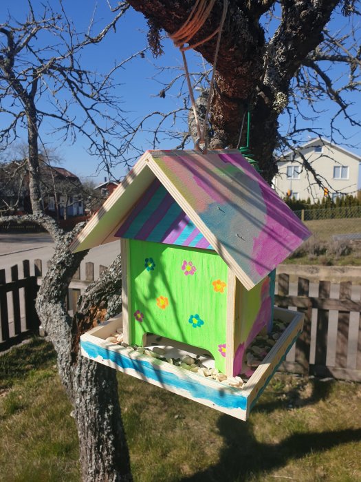 Hemmagjort fågelhus färglagt med randigt mönstrat tak och gröna väggar med handmålade blommor, hängande från trädet.