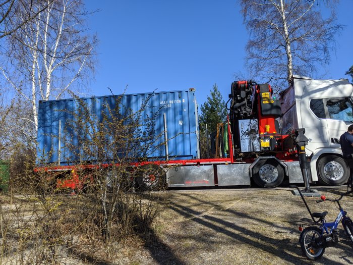 Blå fraktcontainer på röd lastbilsflak med kran, framför träd och barnets cykel.