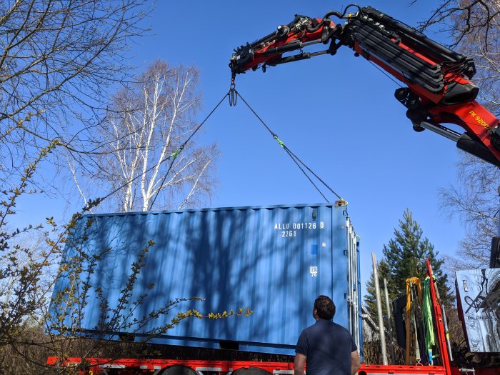 Blå fraktcontainer lyfts av en kranbil medan en person tittar på.