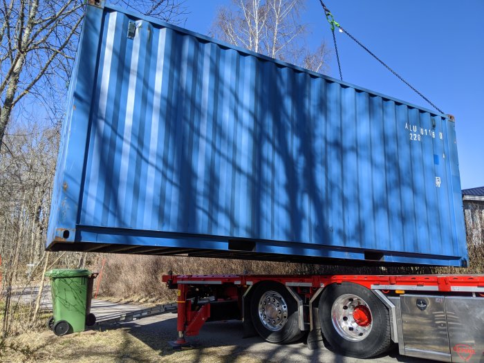 Blå fraktcontainer som lyfts av en lastbil, bredvid en grön soptunna, i solljus med träd i bakgrunden.