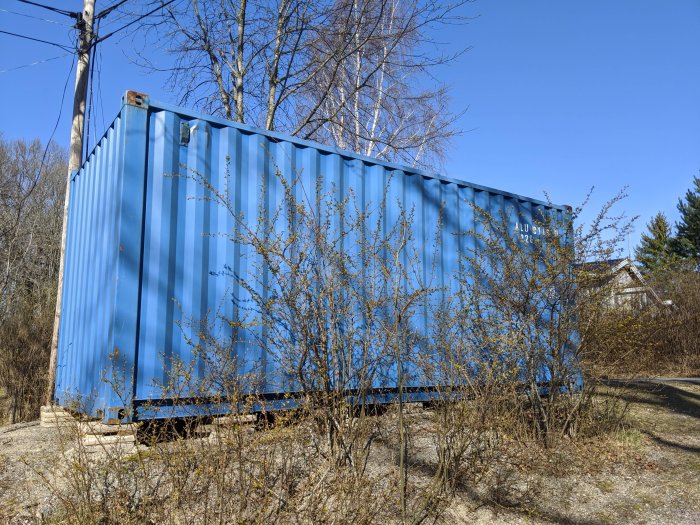 Blå fraktcontainer i en trädgård, tänkt att ersätta gammal snickarbod.