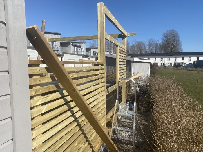 Påbörjad konstruktion av trästaket vid trädgård, verktygsstege synlig, i soligt bostadsområde.