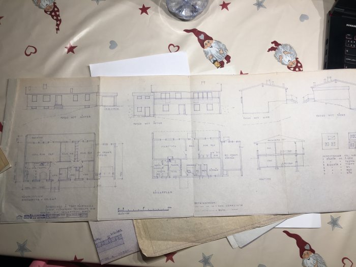 Arkitektritningar utlagda på ett bord med duk med hjärt- och stjärnmönster som visar olika vyer och planlösningar för en byggnad.