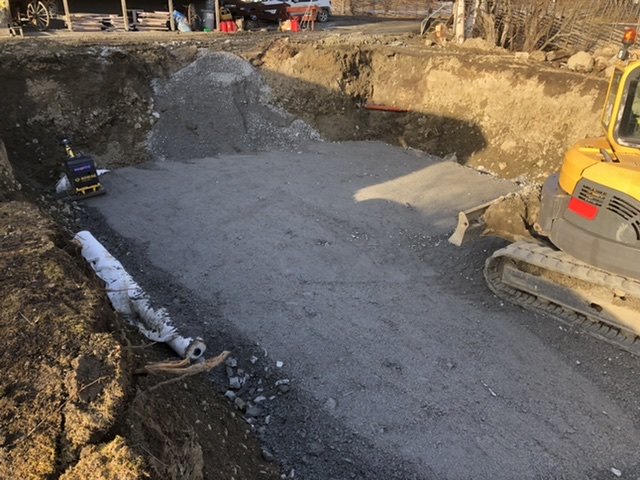 Grävd byggarbetsplats med en hög grus, en gul grävmaskin och byggmaterial synliga i det tidiga byggskedet.