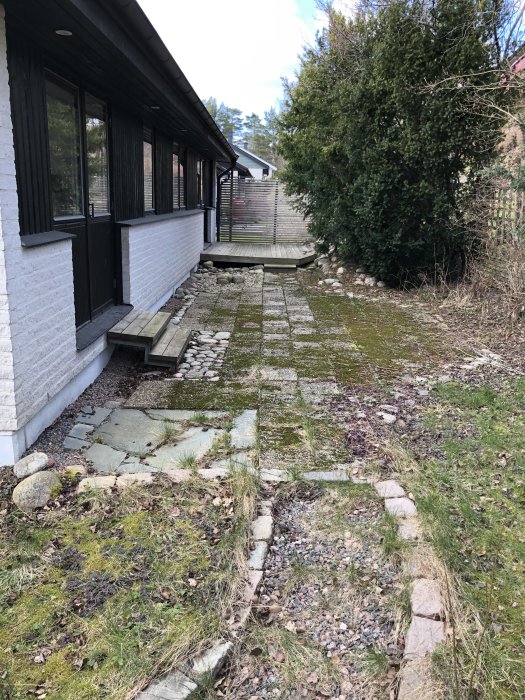 Ett slitet trädäck och gammal stenläggning längs ett hus, omgivet av grönska och stenar.