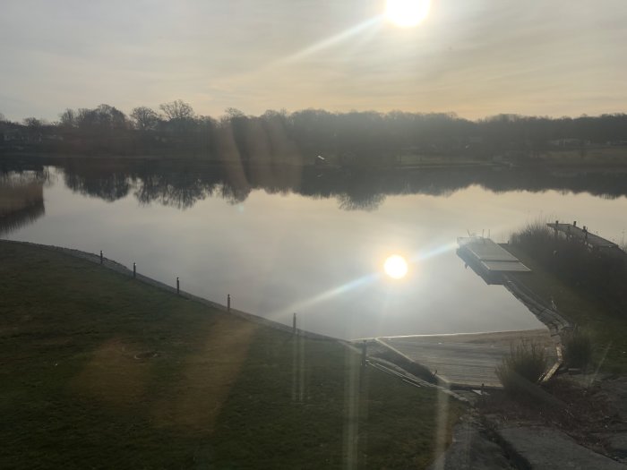Morgonsol skiner över en lugn sjö med en brygga och reflektioner av träd i vattnet, sett från ett fönster.