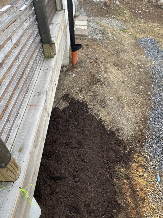 Jord upphögd längs trätrallkanten bredvid plintar och ett hus, med en spade lutad mot väggen.