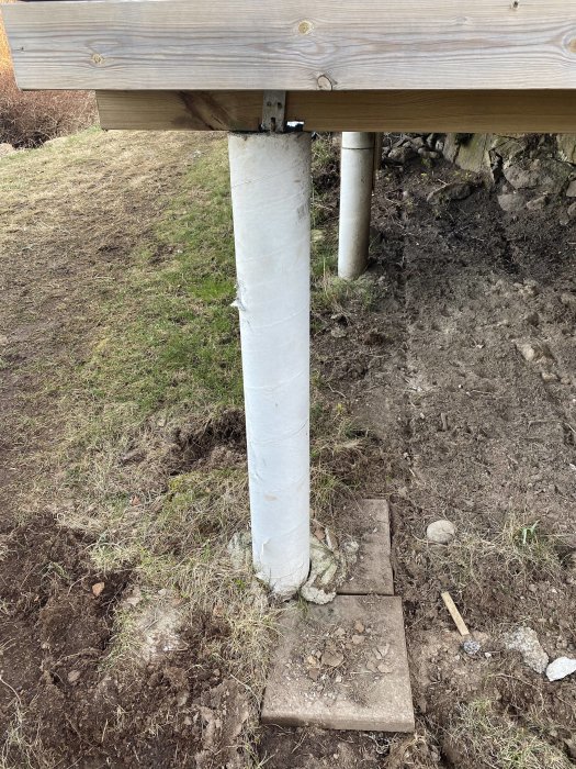 Betongplint under trädäck med omgivande jord bredvid stenplattor, fråga om lämplighet för plantering.