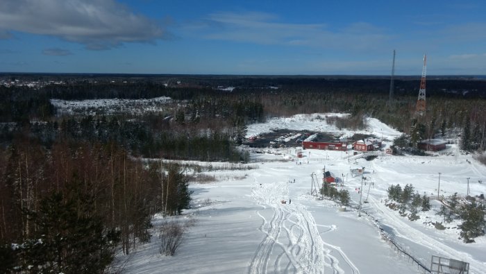 Vy över ett snötäckt landskap med byggnader och vägar, träd i förgrunden, och en klar himmel.