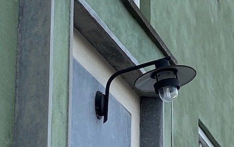 Utomhusvägglampa i svart med genomskinligt glas och skärm, installerad på en husvägg.