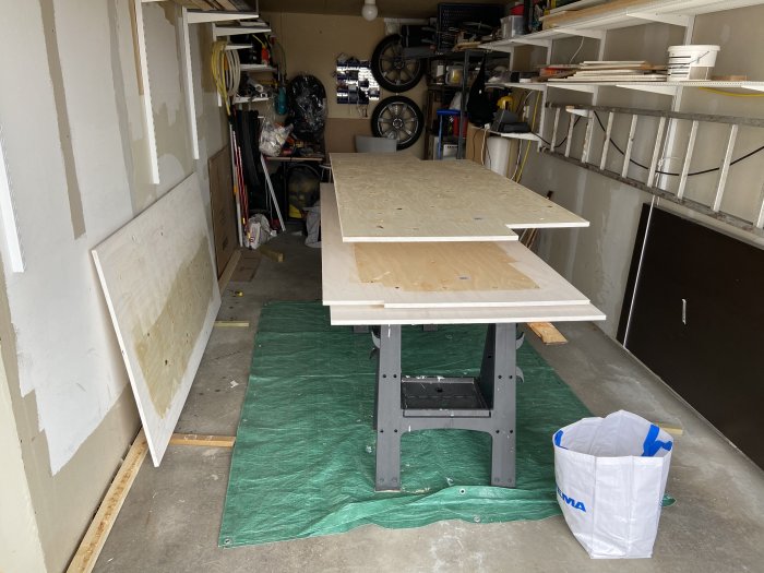 Plywoodskivor på arbetsbockar i ett garage med verktyg och byggmaterial runtomkring.