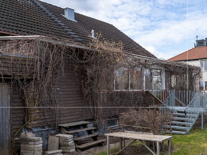 Baksida av hus med brunt trä och uterum täckt av lövverk, omgiven av en trappa och terrass.