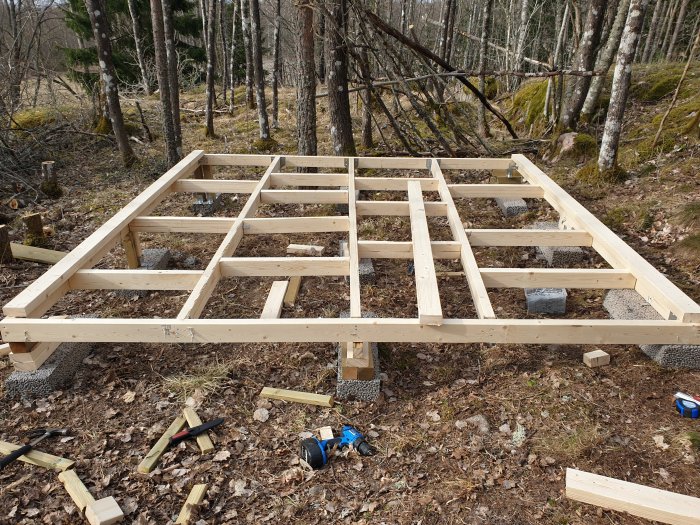 Nästan färdigställd golvstomme av 45x95 dimensioner i en skogsmiljö, verktyg och träbitar syns på marken.