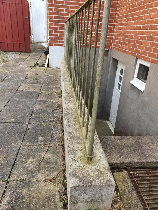 Smutsiga betongplattor och en betongmur med påväxt nära ett rött tegelhus med vita detaljer och en vit dörr.