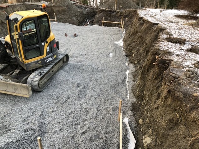 Grävmaskin på singelbädd bredvid öppen grävning för dränering omgiven av lera.