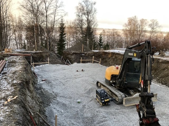 Byggarbetsplats med dränering och singelbädd, grävmaskin till vänster, omgiven av lera och natur.