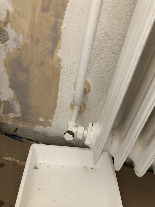 Ett gammalt radiator-element med öppet mässingsanslutning och en uppsamlingskärl under, mot avskalad väggbakgrund.
