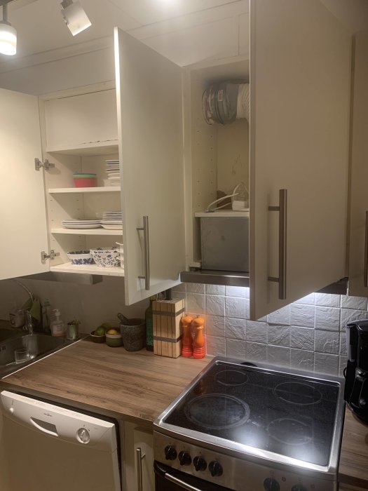Kök med öppet skåp som visar befintlig fläktkanal, spis och bänkskiva samt delvis dold diskho.