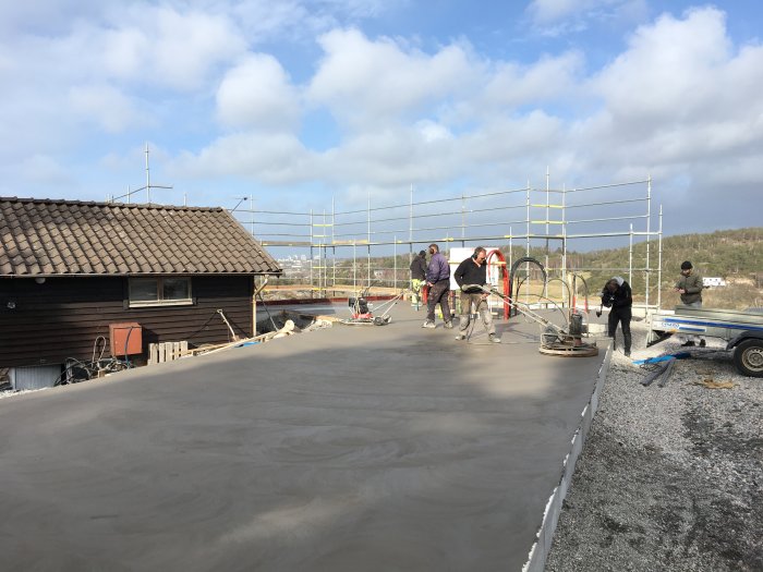 Arbetslag gjuter betongplatta på husbygge med byggnadsställning i bakgrunden och klarblå himmel ovan.