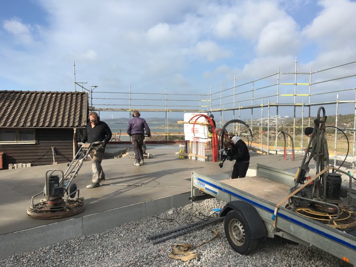 Byggplats med arbetare som slipar betong, ställningar och havsutsikt i bakgrunden.