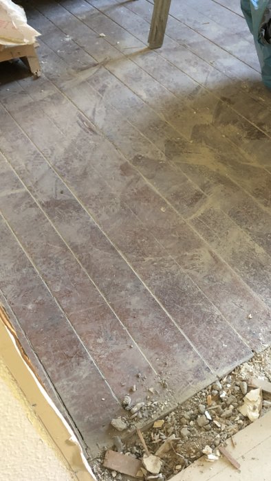 Ett gammalt trägolv med breda plankor täckt av damm och smuts under renovering, med synliga spikar och stenar.