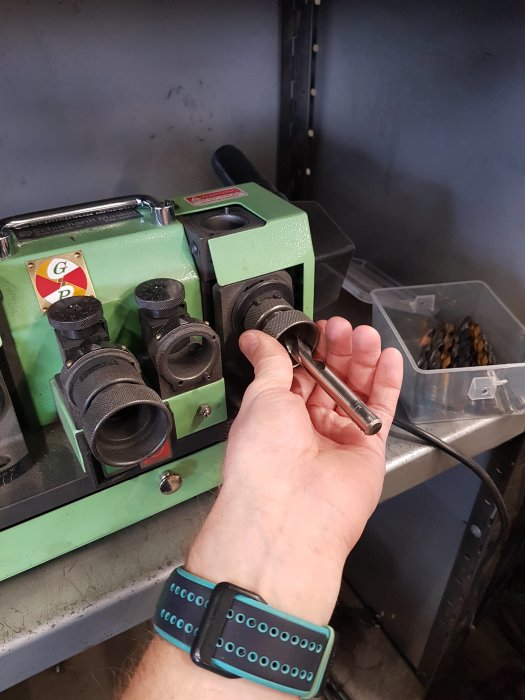 Hand håller en slipad del framför en bordsslipmaskin på en arbetsbänk.