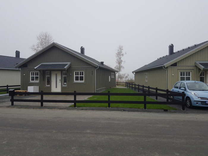 Svart staket med 90x90 stolpar framför ett grönt gräsmatta och hus, dimmig bakgrund.