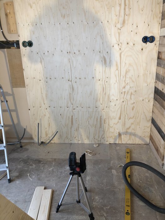Plywoodvägg med uttag och nivelleringslaser på stativ under konstruktion.